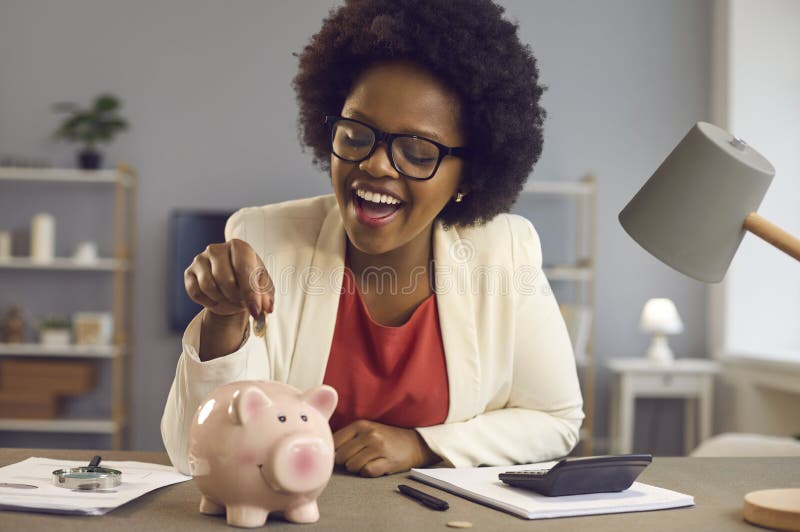 Happy black businesswoman puts money in piggy bank sitting at office desk with calculator