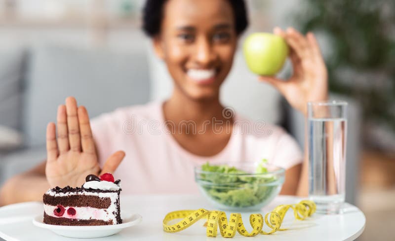 Happy black woman choosing fresh apple over unhealthy cake, refusing to eat dessert at home, selective focus