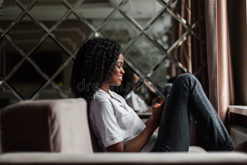 Happy black woman sitting on the sofa and holding a phone. Woman typing a messege.