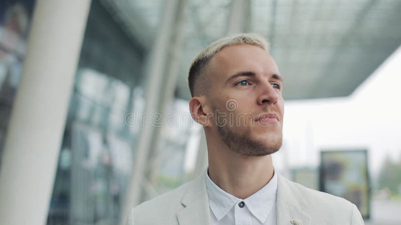 Happy blond attractive businessman walking outside near big office building and looking around. Man looking satisfied