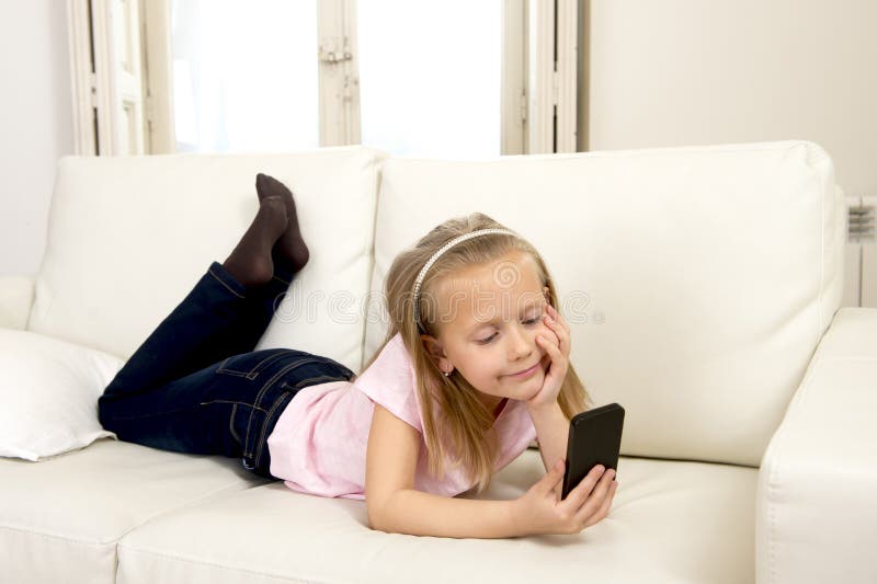 Happy blond little girl on home sofa using internet app on mobile phone