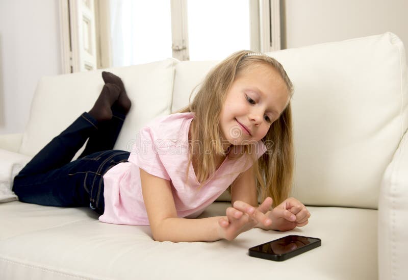 Happy blond little girl on home sofa using internet app on mobile phone