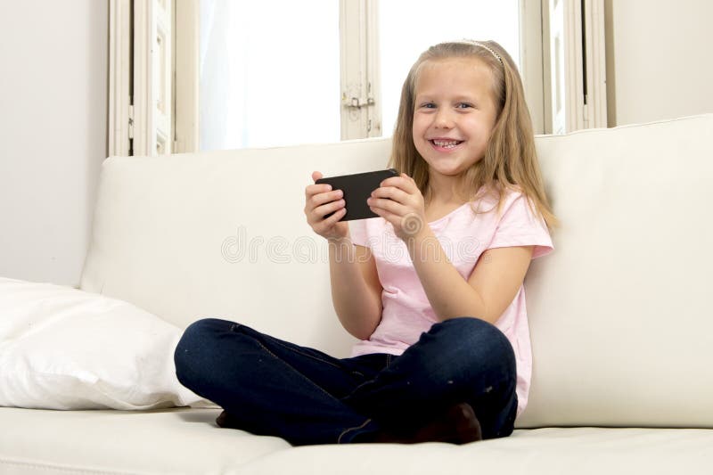 Happy blond little girl on home sofa using internet app on mobile phone