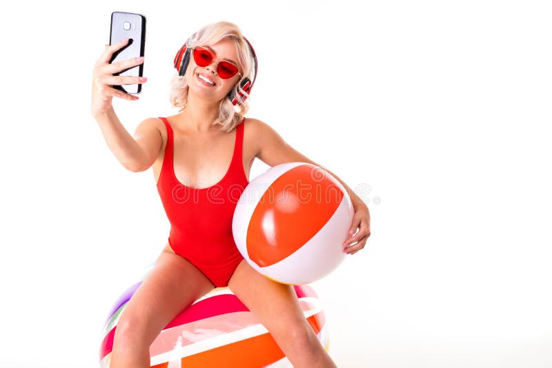 Happy blonde caucasian female lies in swimsuit with big rubber mattress and colourful ball, earphones and smiles