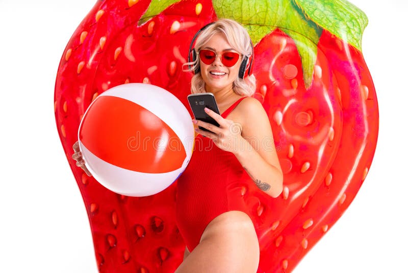 Happy blonde caucasian female lies in swimsuit with big rubber mattress and colourful ball, earphones and smiles