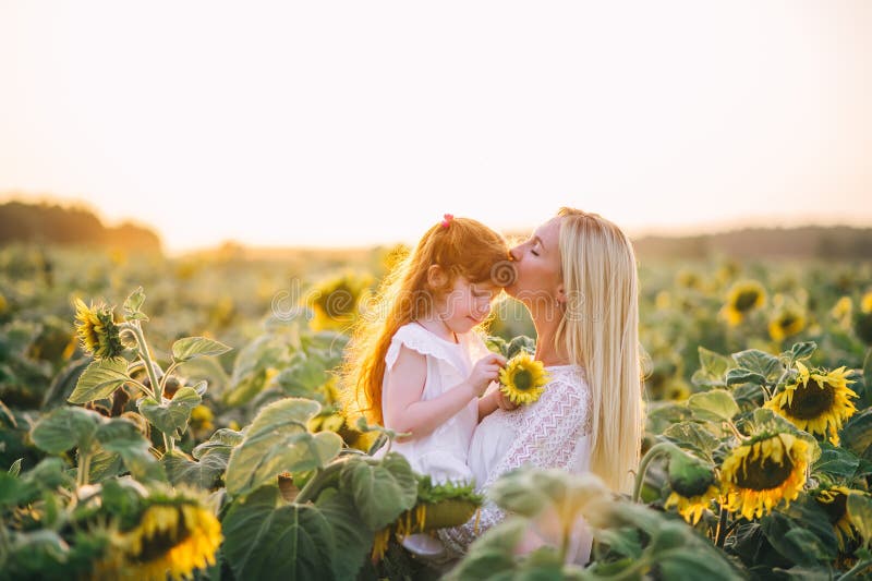 Happy mother kisses her little red-heired daughter in a field with yellow sunflowers on sunset