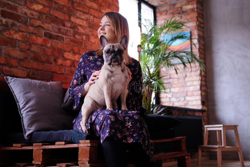 Happy blonde woman in dress sitting with her cute pug on a handmade sofa in room with loft interior. stock photo Happy blonde woman in dress sitting with her cute pug on a handmade sofa in room with loft interior. stock photo