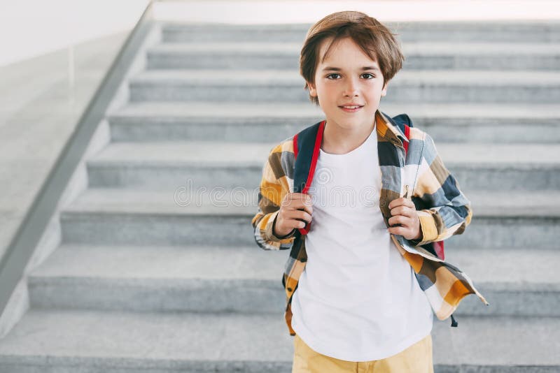 A happy boy with a backpack stands on the steps in front of the entrance to the school and smiles beautifully. Beginning of the