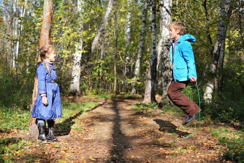 Happy boy jumps with skipping rope and girl looks at him in forest on sunny day.