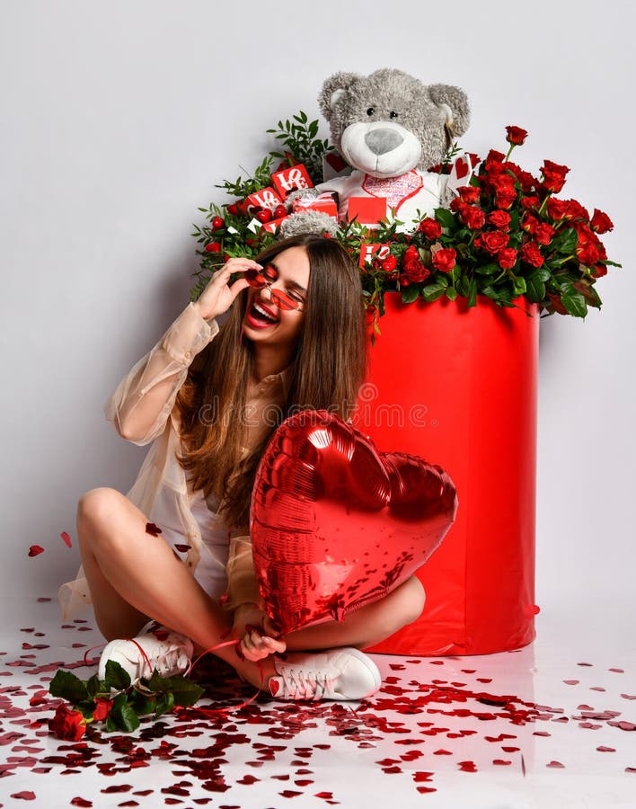 Happy laughing young brunette woman celebrate valentines day or birthday party is sitting at gift box with flowers roses under confetti falling holding red heart balloon on white. Happy laughing young brunette woman celebrate valentines day or birthday party is sitting at gift box with flowers roses under confetti falling holding red heart balloon on white