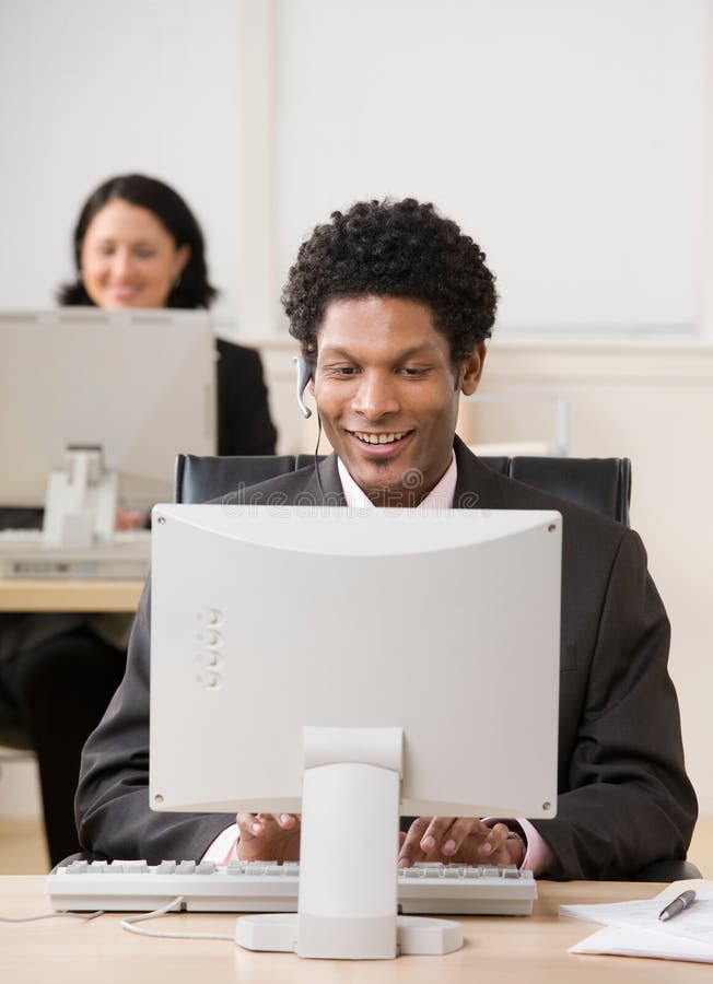 Happy businessman in headset working on computer with co-worker in background