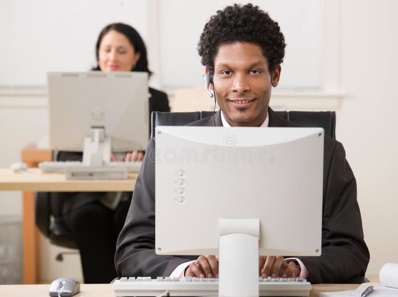 Happy businessman in headset working on computer with co-worker in background