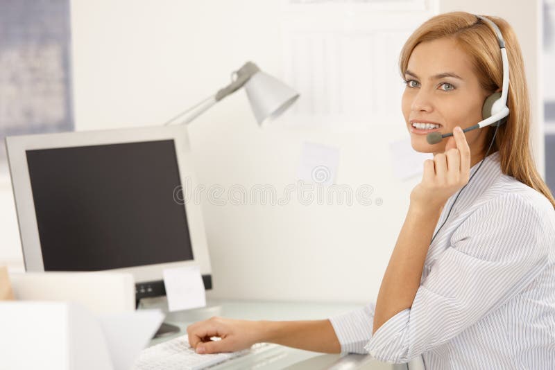 Happy call center worker girl sitting at desk in office, using desktop computer with headset, smiling. Happy call center worker girl sitting at desk in office, using desktop computer with headset, smiling.