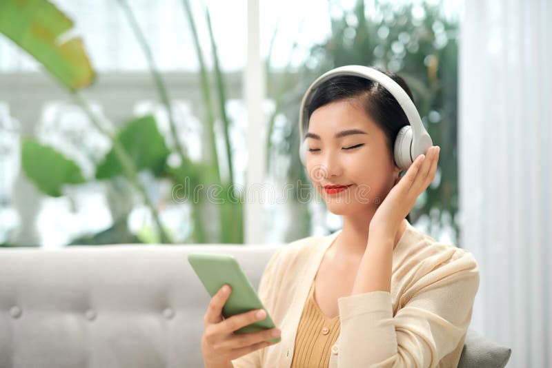 Happy carefree asian girl wearing wireless headphones listening to music with eyes closed sit on sofa