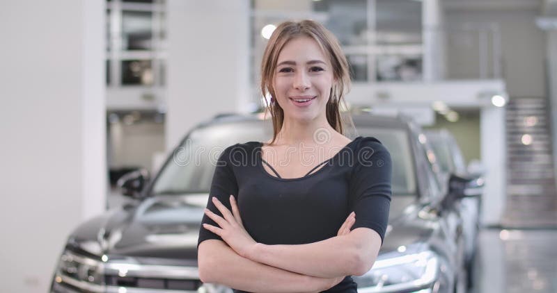 Happy Caucasian girl posing at the background of blurred black automobile. Young smiling woman buying or leesing car in