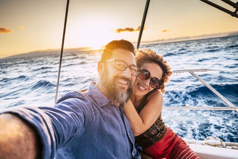 Happy and cheerful people enjoying the travel and trip on a sail boat with ocean and sunset sunlight in background - traveler lifestyle for adult men and women smiling doing a selfie picture