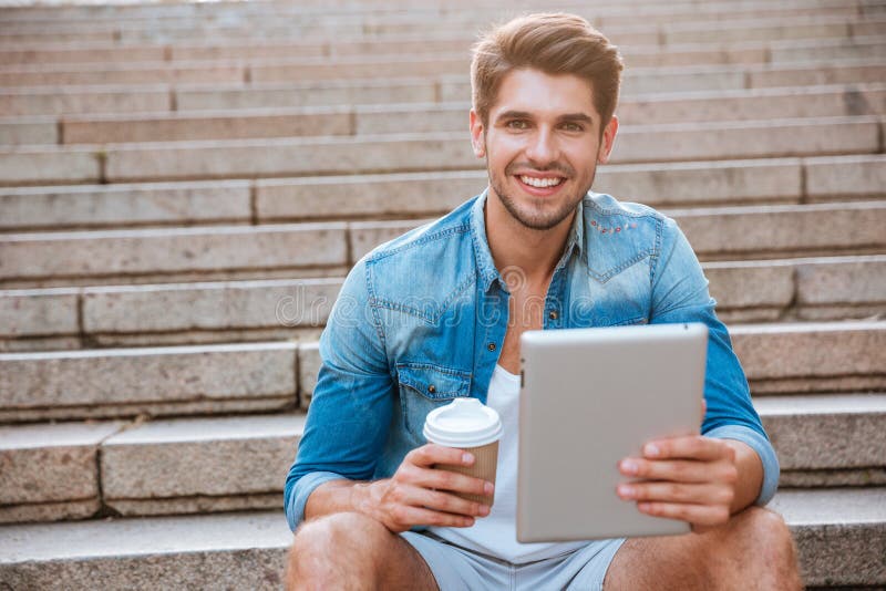 Happy cheerful student with tablet and coffee cup sitting outdoors