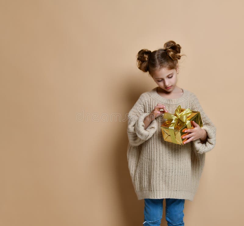 Happy child girl with gift box. Portrait of a little girl in a warm nude knitted sweater on a beige background. A child holds a gold box with a gift at arms. The stock image Happy child girl with gift box. Portrait of a little girl in a warm nude knitted sweater on a beige background. A child holds a gold box with a gift at arms. The stock image