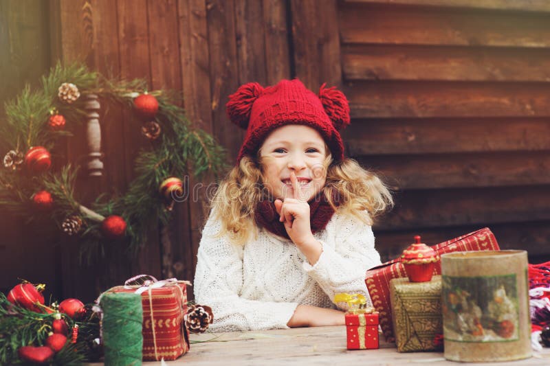 Happy child girl in red hat and scarf wrapping Christmas gifts at cozy country house, decorated for New Year and Christmas