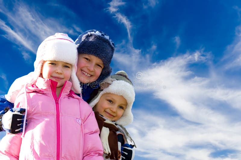 Tre felici, bambini sorridenti in posa per una foto vestita in inverno cappotti e cappelli contro un cielo blu brillante con ciuffi di nuvole bianche.