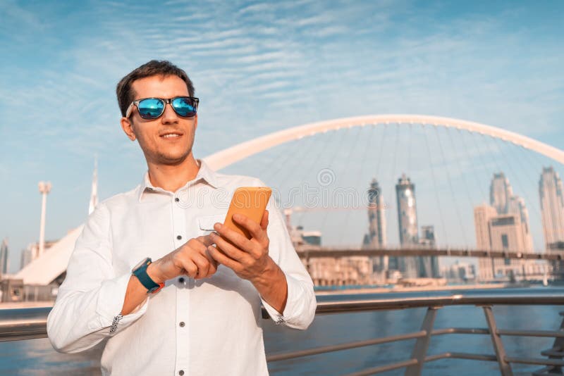 Happy young guy smiles and uses his mobile phone against the background of the wonderful modern architecture of the royalty free stock photo