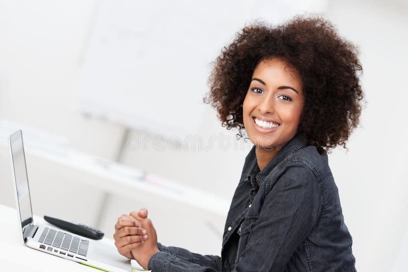 Happy contented young African American woman with an afro hairstyle sitting at her desk in front of a laptop. Happy contented young African American woman with an afro hairstyle sitting at her desk in front of a laptop