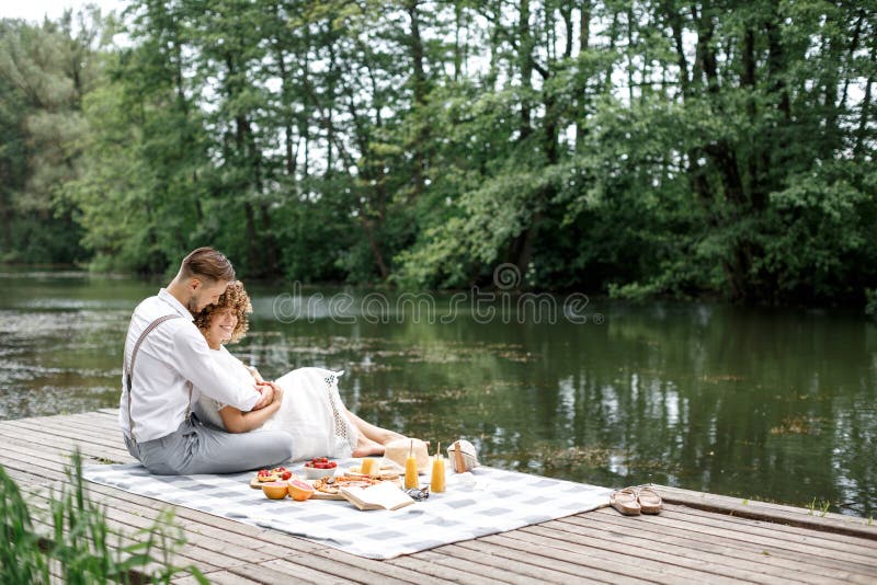 Happy couple have a rest in the park near the lake on a picnic. A couple sits on a plaid blanket on a picnic near a meal