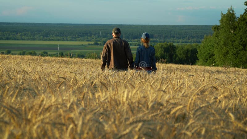 Happy couple, middle-aged man and woman talking and smiling while walking on wheat field at sunset summer evening.