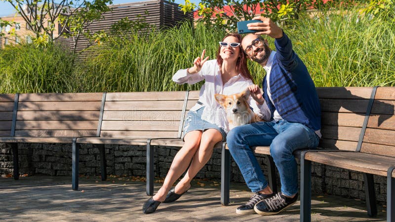 The happy couple are sitting on the bench in the park with little dog and do selfie portrait. Cute corgi Pembroke puppy on its