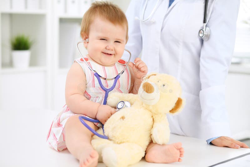 Happy cute baby at health exam at doctor`s office. Toddler girl is sitting and keeping stethoscope and teddy bear