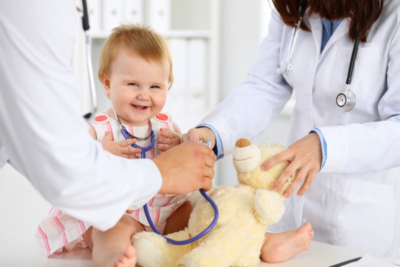 Happy cute baby at health exam at doctor`s office. Toddler girl is sitting and keeping stethoscope and teddy bear. Nurse, care.