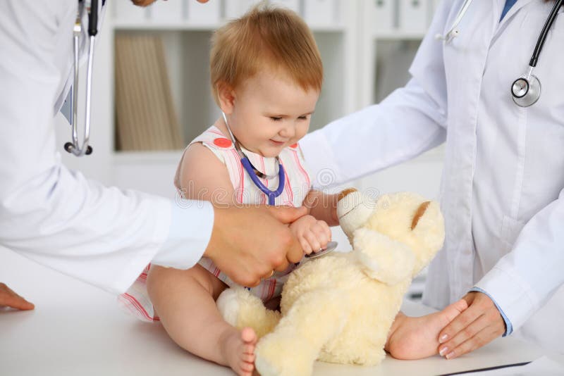 Happy cute baby at health exam at doctor`s office. Toddler girl is sitting and keeping stethoscope and teddy bear. Holding, hospital.