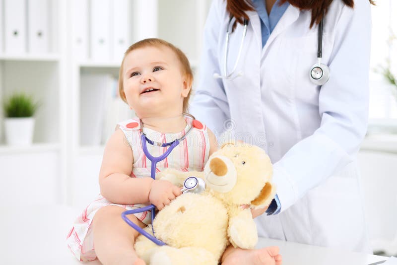 Happy cute baby at health exam at doctor`s office. Toddler girl is sitting and keeping stethoscope and teddy bear. Clinician, caucasian.