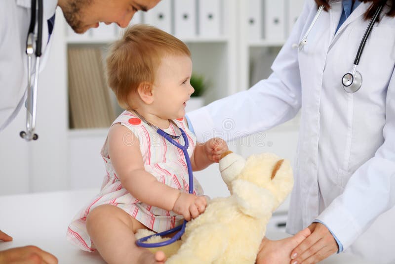 Happy cute baby at health exam at doctor`s office. Toddler girl is sitting and keeping stethoscope and teddy bear. Medicine, medical.