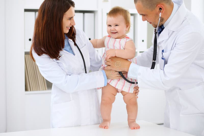 Happy cute baby at health exam at doctor`s office. Toddler girl is standing while have been keeping by two doctors