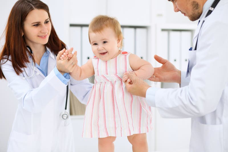 Happy cute baby at health exam at doctor`s office. Toddler girl is standing while have been keeping by two doctors