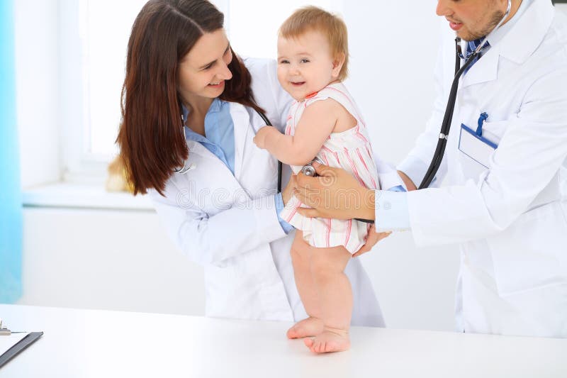 Happy cute baby at health exam at doctor`s office. Toddler girl is standing while have been keeping by two doctors. Medical, medicine.