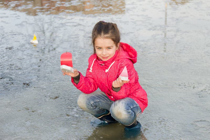 Happy cute little girl in rain boots playing with handmade colorful ships in the spring creek standing in water