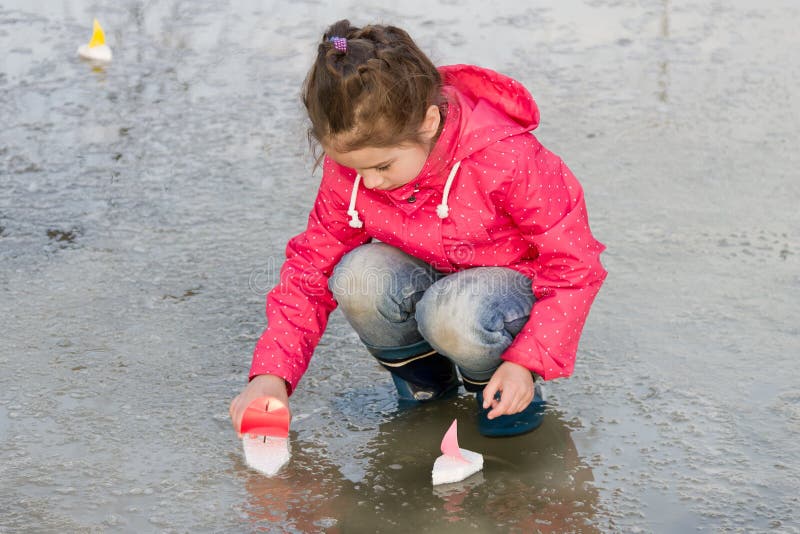 Happy cute little girl in rain boots playing with handmade colorful ships in the spring water puddle