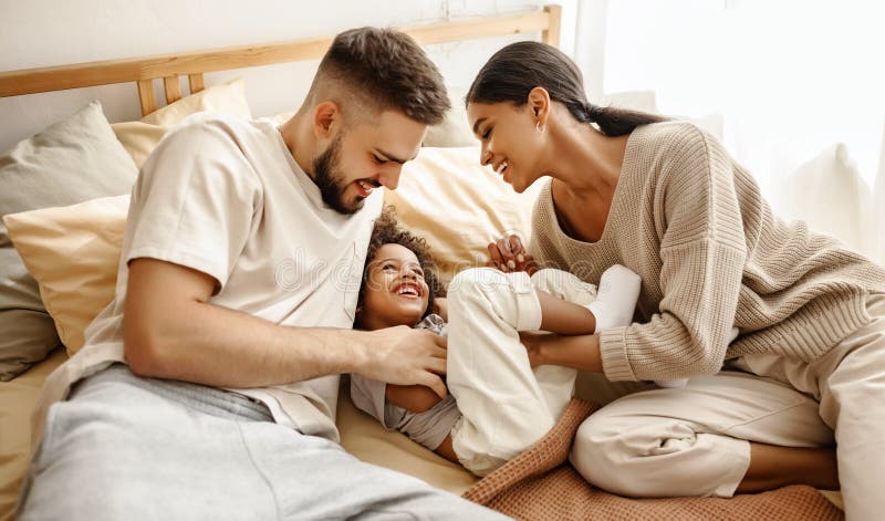 Happy diverse family mom, dad and child  laughing, playing and tickles   in bed   at home