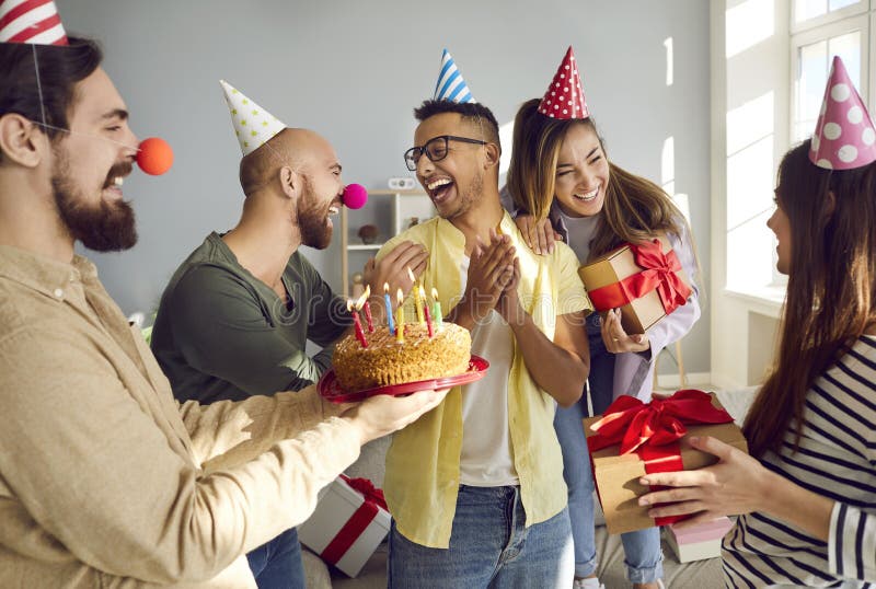 Caring diverse friends greeting excited mate with happy birthday. Smiling multiethnic young people congratulate overjoyed colleague with present and cake with candles. Anniversary concept. Caring diverse friends greeting excited mate with happy birthday. Smiling multiethnic young people congratulate overjoyed colleague with present and cake with candles. Anniversary concept.