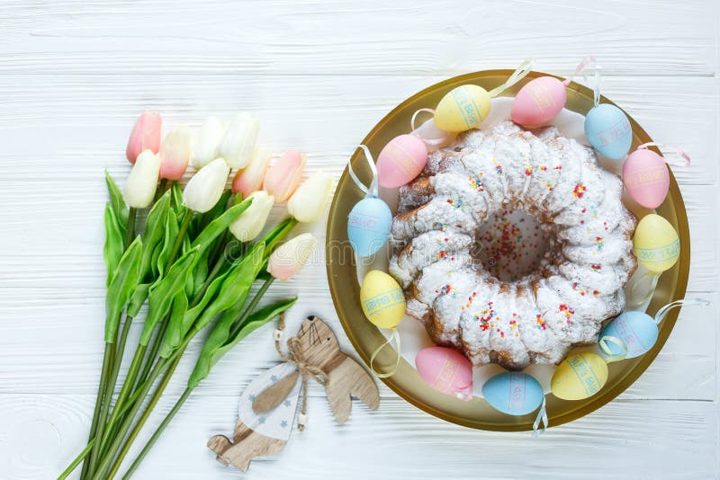 Happy Easter! Golden tray with plate wit cake and hand painted colorful eggs, tulips on white wooden table. Close up. Decoration