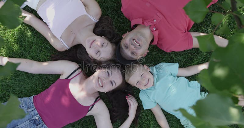 Happy Family Laying Down on a Green Grass and Looking up Smiling in a Shade of a Tree in the Summer Top View
