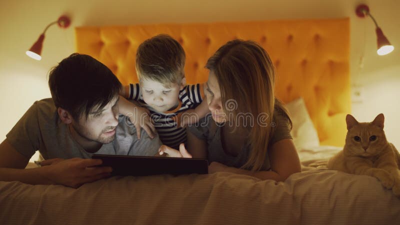 Happy family with little son and funny cat lying in bed at home and using tablet computer for playing game before stock image Happy family with little son and funny cat lying in bed at home and using tablet computer for playing game before stock image