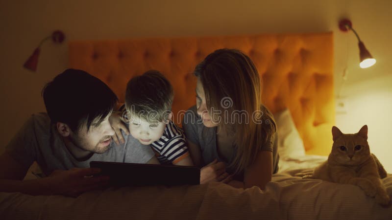 Happy family with little son and funny cat lying in bed at home and using tablet computer for watching cartoon movie