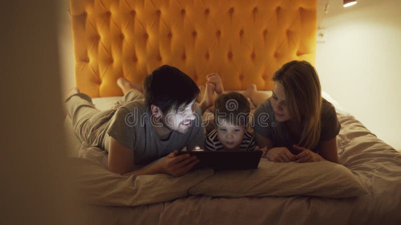 Happy family with little son a lying in bed at home and using tablet computer for watching cartoon movie before sleeping
