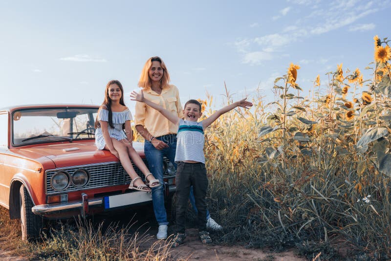 Happy family near sunflowers field, mom with two children, girl teen in dress sitting on hood of the red retro car