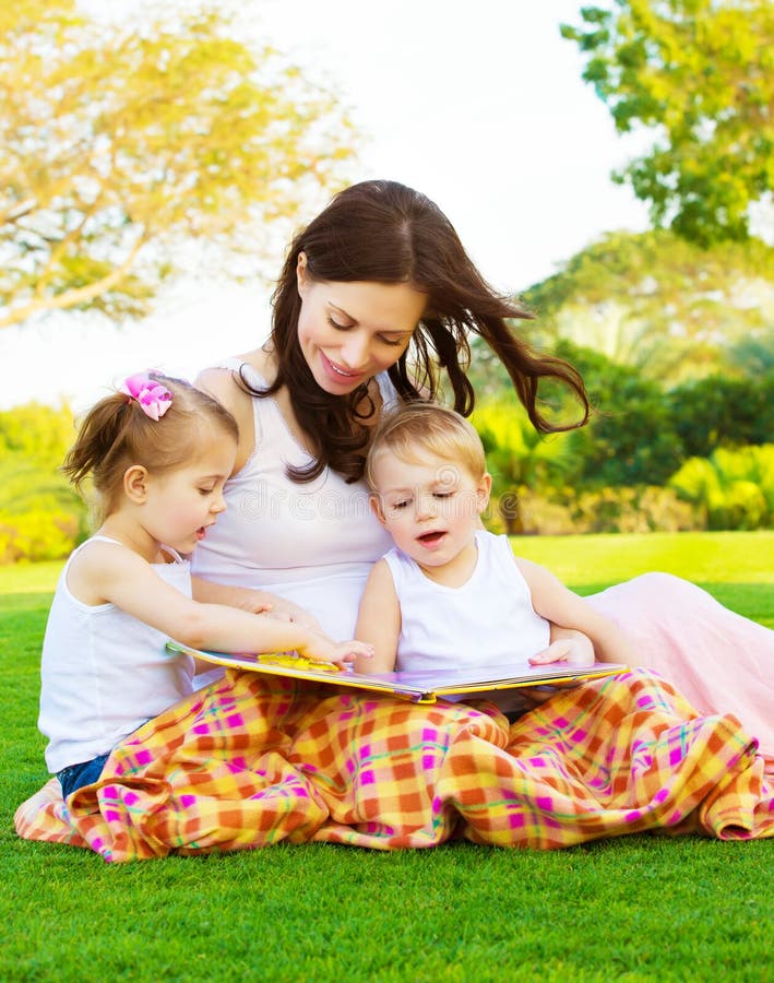 Picture of beautiful women with daughter and son sitting down on green grass field and read fairytale, cheerful mother and two pretty kids enjoying book on backyard in spring, happy family concept. Picture of beautiful women with daughter and son sitting down on green grass field and read fairytale, cheerful mother and two pretty kids enjoying book on backyard in spring, happy family concept