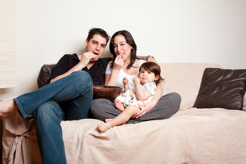 Beautiful happy family, father mother toddler daughter child, eating snacks while sitting on sofa couch in livingroom. Beautiful happy family, father mother toddler daughter child, eating snacks while sitting on sofa couch in livingroom.