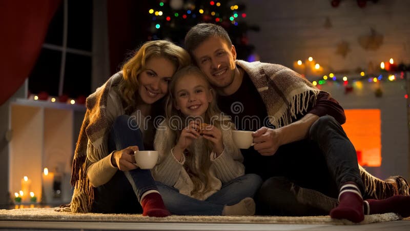 Happy family of three drinking hot cocoa near Christmas tree, miraculous day stock photos Happy family of three drinking hot cocoa near Christmas tree, miraculous day stock photos